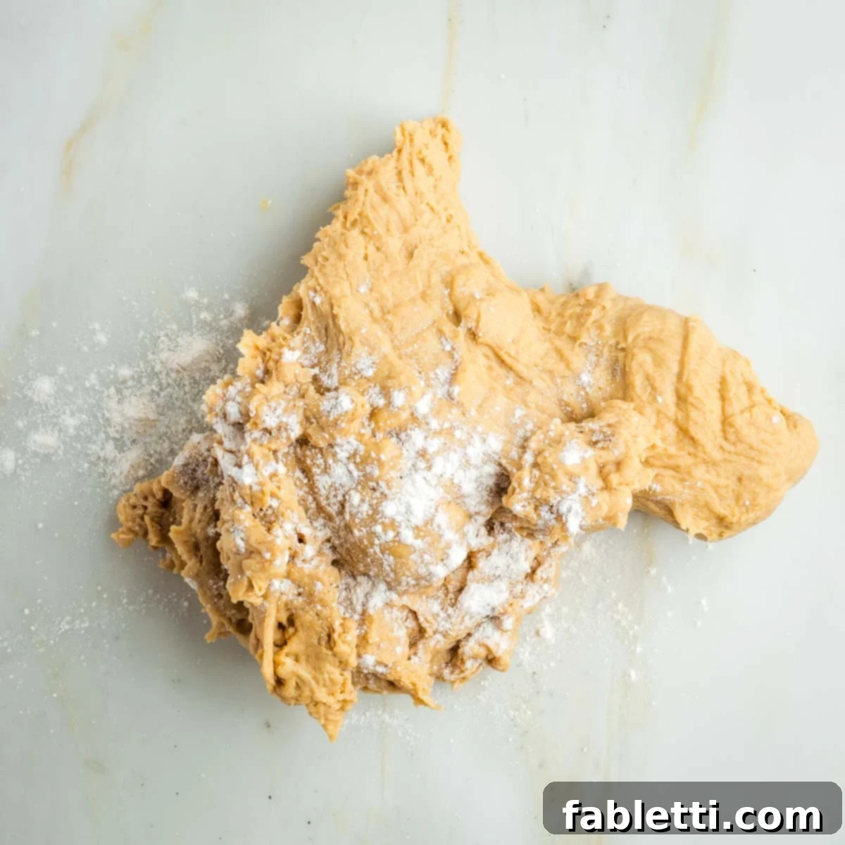 Pillowy Sweet Challah Crown 17 A mound of freshly kneaded bread dough rests on a marble counter, lightly dusted with flour, ready for shaping.