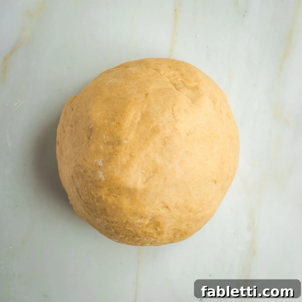 Pillowy Sweet Challah Crown 18 A perfectly smooth, round ball of challah dough rests on a clean marble countertop, signifying it's ready for its first rise.