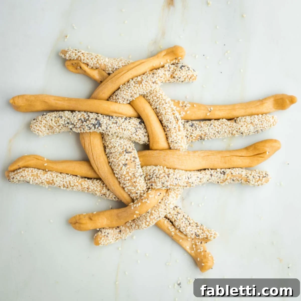 Pillowy Sweet Challah Crown 28 Groups of challah dough strands, some with seeds and some plain, intricately crossing over and under each other in a patterned weave, forming the start of a round braid.