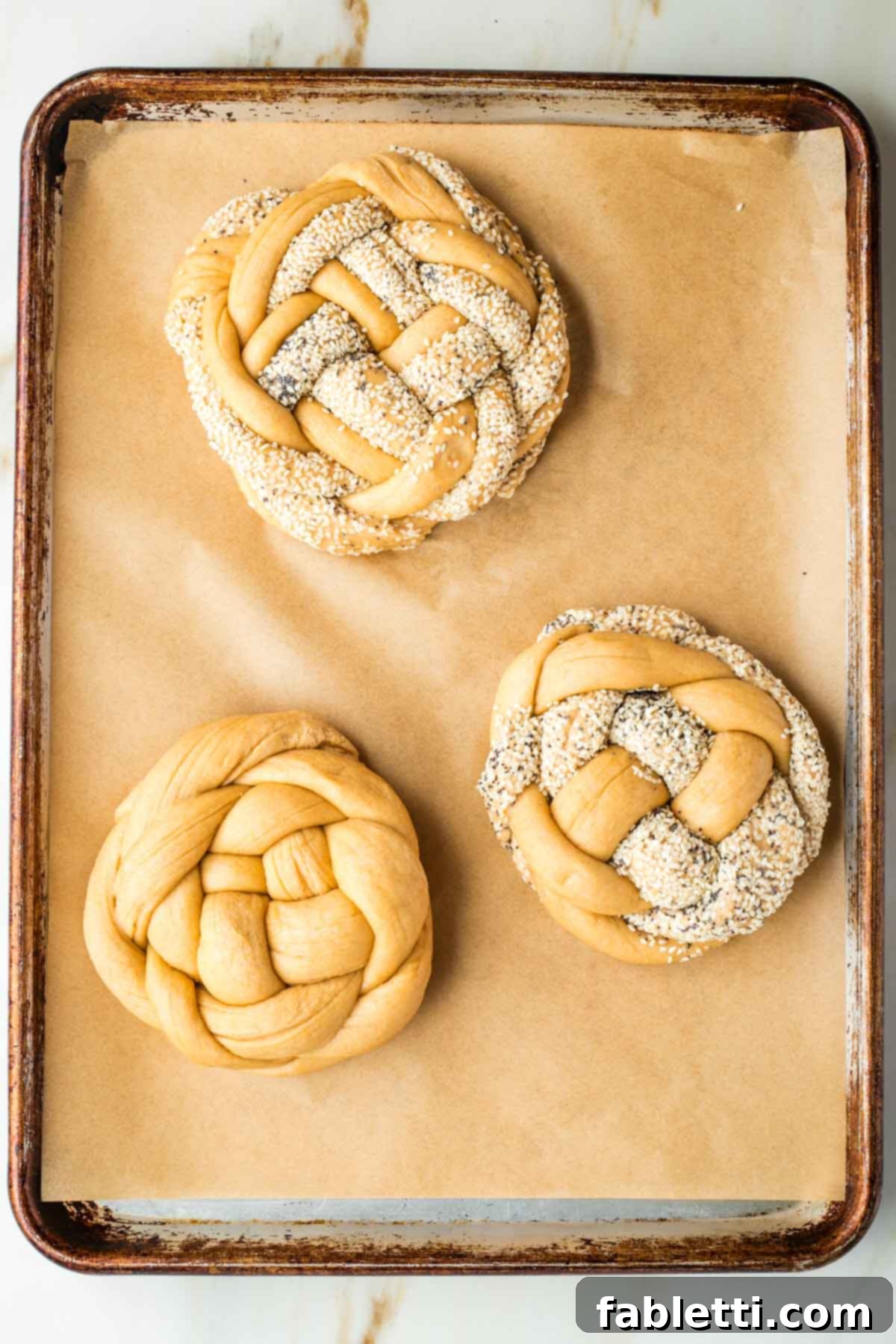 Pillowy Sweet Challah Crown 39 Three beautifully braided round loaves of challah bread, some adorned with poppy and sesame seeds, neatly arranged on a parchment-lined baking sheet, prepared for their second rise.
