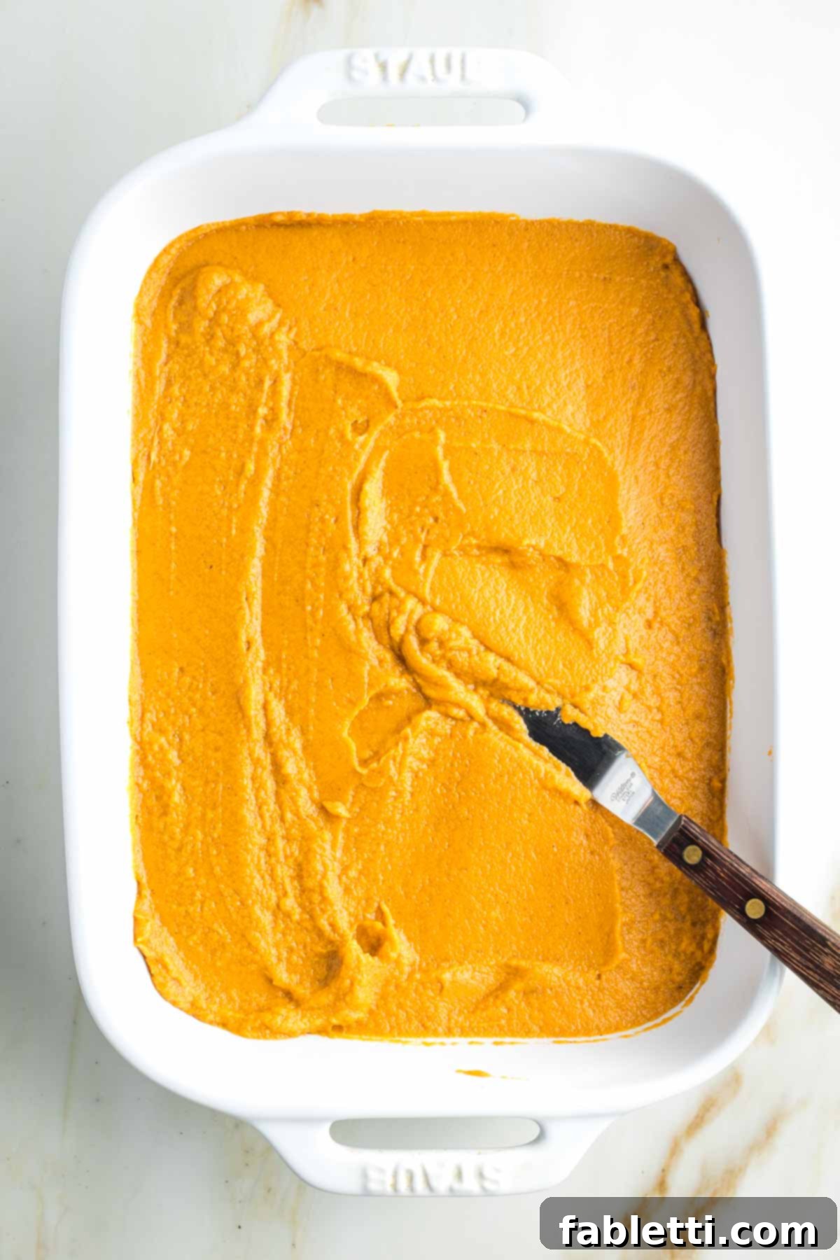 Golden Maple Pecan Sweet Potato Casserole 15 An offset spatula spreading a layer of pureed sweet potatoes evenly into a white baking dish, creating a smooth base.