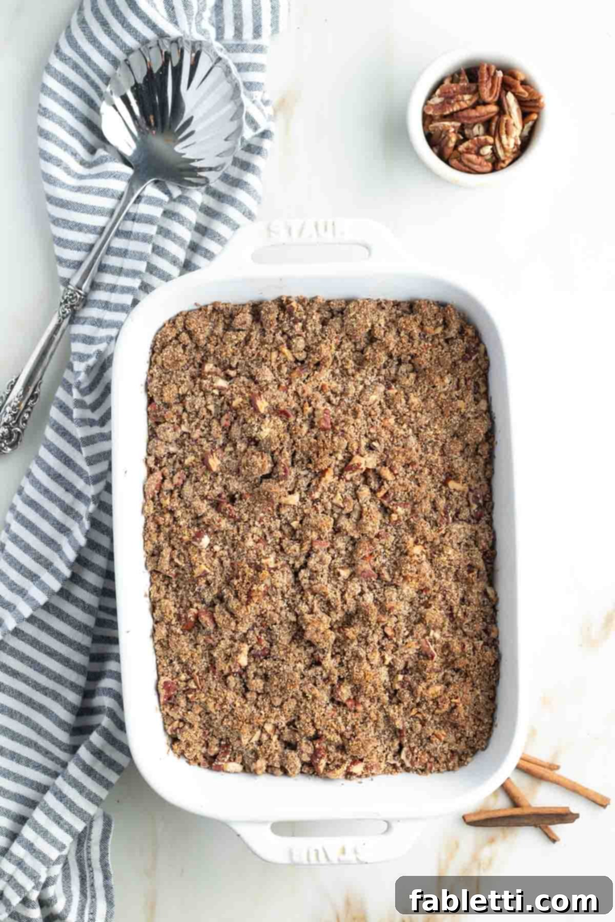 Golden Maple Pecan Sweet Potato Casserole 17 Sweet potato casserole with crumbly pecan topping in a baking dish, styled photo with extra pecans, a cinnamon stick, and a serving spoon, ready to be served.