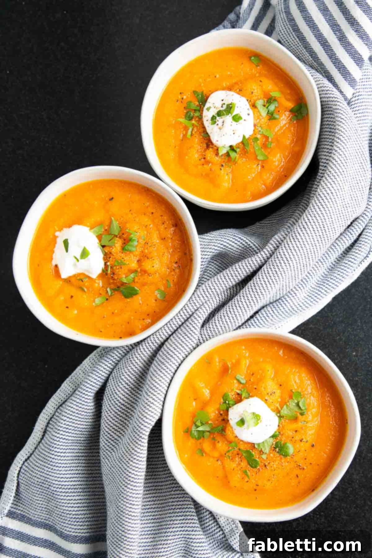 Golden Vegan Carrot Ginger Soup 16 Three individual bowls filled with vibrant orange carrot soup, each delicately topped with a dollop of creamy vegan sour cream and a scattering of fresh herbs.
