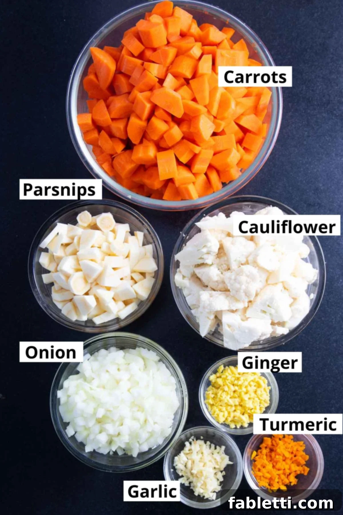 Golden Vegan Carrot Ginger Soup 7 A neat arrangement of various chopped and minced vegetables, clearly labeled as carrots, parsnips, cauliflower, onions, ginger, turmeric, and garlic, ready for cooking.
