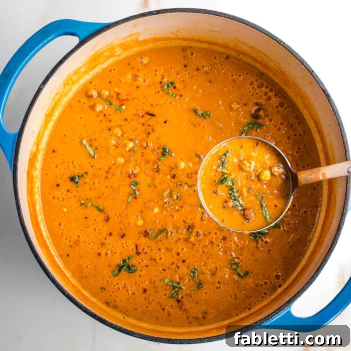 Creamy Vegan Tomato Soup 11 Dutch oven with creamy tomato soup on a ladle, garnished with fresh basil, crushed red pepper, and crunchy chickpeas.