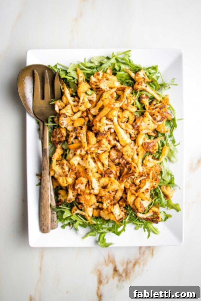 Large white rectangular platter, scattered with arugula and then topped with paprika roasted cauliflower, with bronze serving pieces on the left side of plate. 