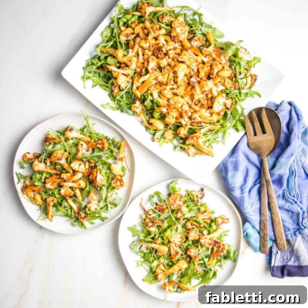 Large rectangular plate filled with arugula and then roasted cauliflower. two circular rimmed plates with same cauliflower salad and bronze serving pieces  off to the side resting on a blue tea towel. 