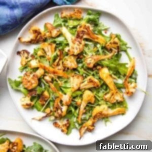 Close up of a white plate, with arugula and roasted cauliflower with paprika. The edge of a blue napkin in the upper left corner.