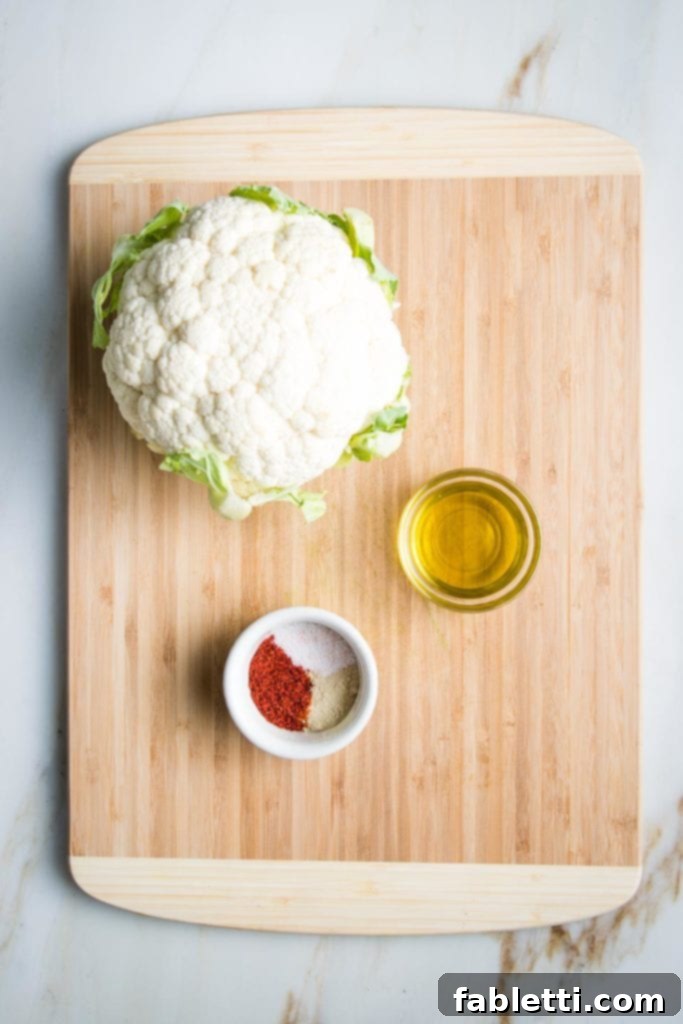 Wooden cutting board with a head of cauliflower, a small glass dish with olive oil and a small white dish with paprika, salt and white pepper
