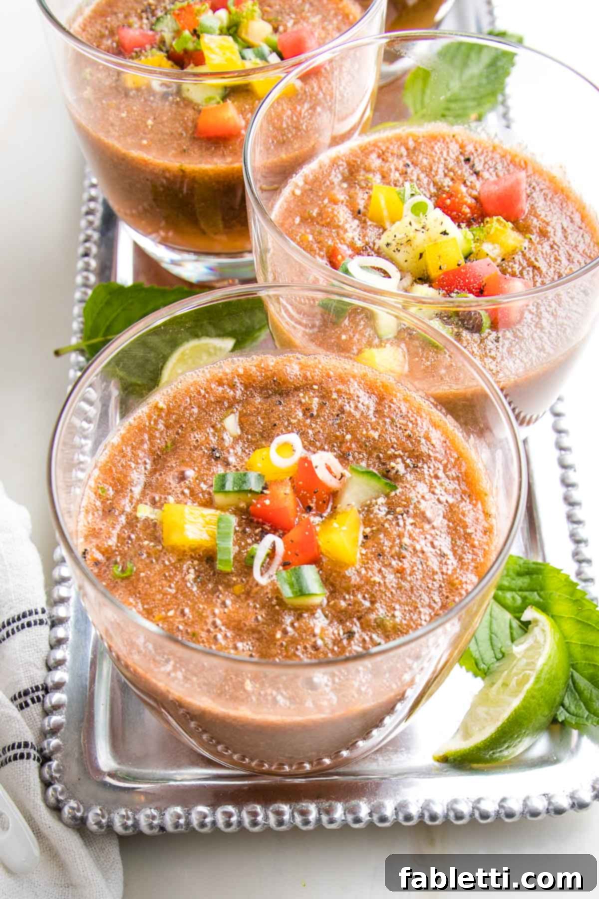 A glistening silver tray presents elegantly portioned glasses of Watermelon Gazpacho, adorned with a vibrant medley of colorful, diced vegetables gracefully floating atop. Fresh mint sprigs and bright lime wedges artfully arranged on the tray complete this invigorating summer spectacle.