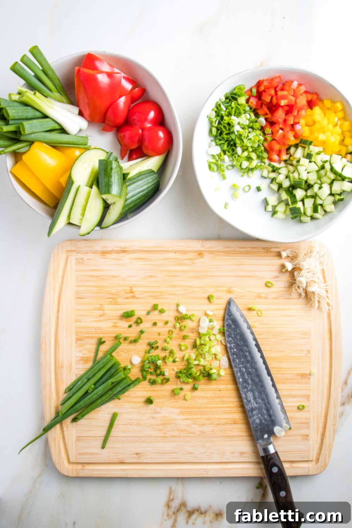 Scallions are being thinly sliced on a wooden cutting board. Above, two white bowls showcase the prepared vegetables: one with chunky bell peppers, scallions, and cucumbers for blending, and the other with the same veggies finely diced for garnish.
