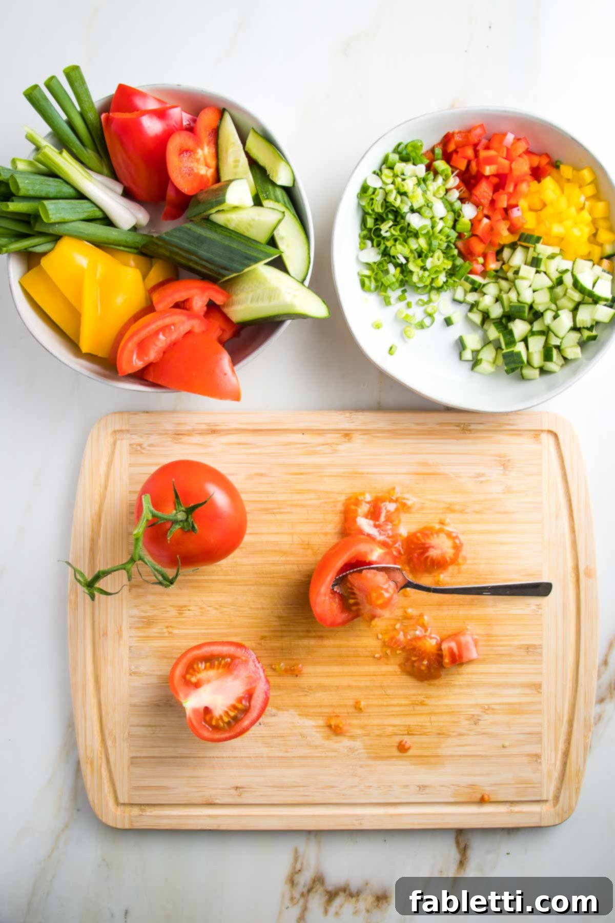 A cutting board displays a tomato on the vine and another that has been halved, with seeds being scooped out using a grapefruit spoon. Above the board are two white bowls: one containing chunks of colorful vegetables for blending, and the other with small diced yellow and red peppers, cucumbers, and scallions for garnish.