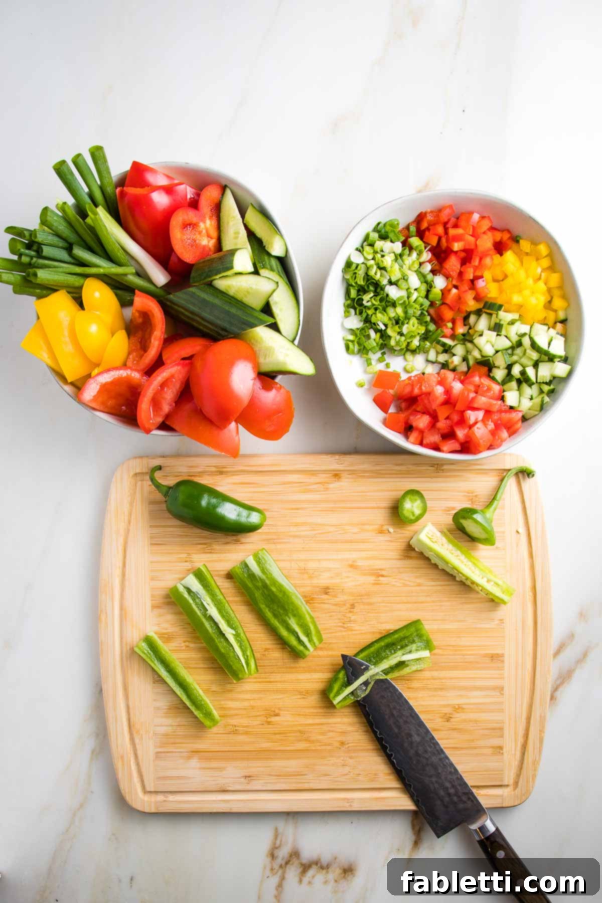 Two bowls are featured: one brimming with chunky vegetables, the other with finely diced versions of the same—red and yellow peppers, cucumbers, scallions, and tomatoes. On the cutting board, a jalapeño is trimmed, and its membranes are being removed from slices.