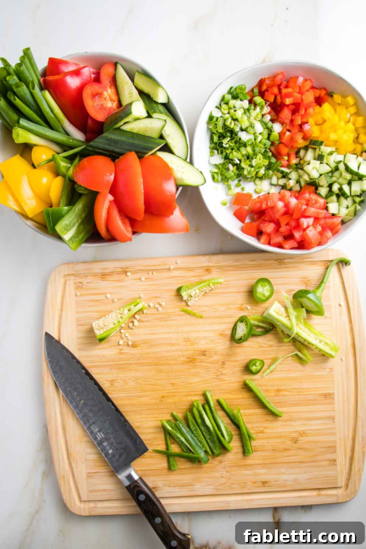 A cutting board shows a trimmed and seeded jalapeño being cut into strips. Above are two bowls: one filled with chunky vegetables, the other with small diced versions of the same—red and yellow peppers, scallions, and cucumbers.