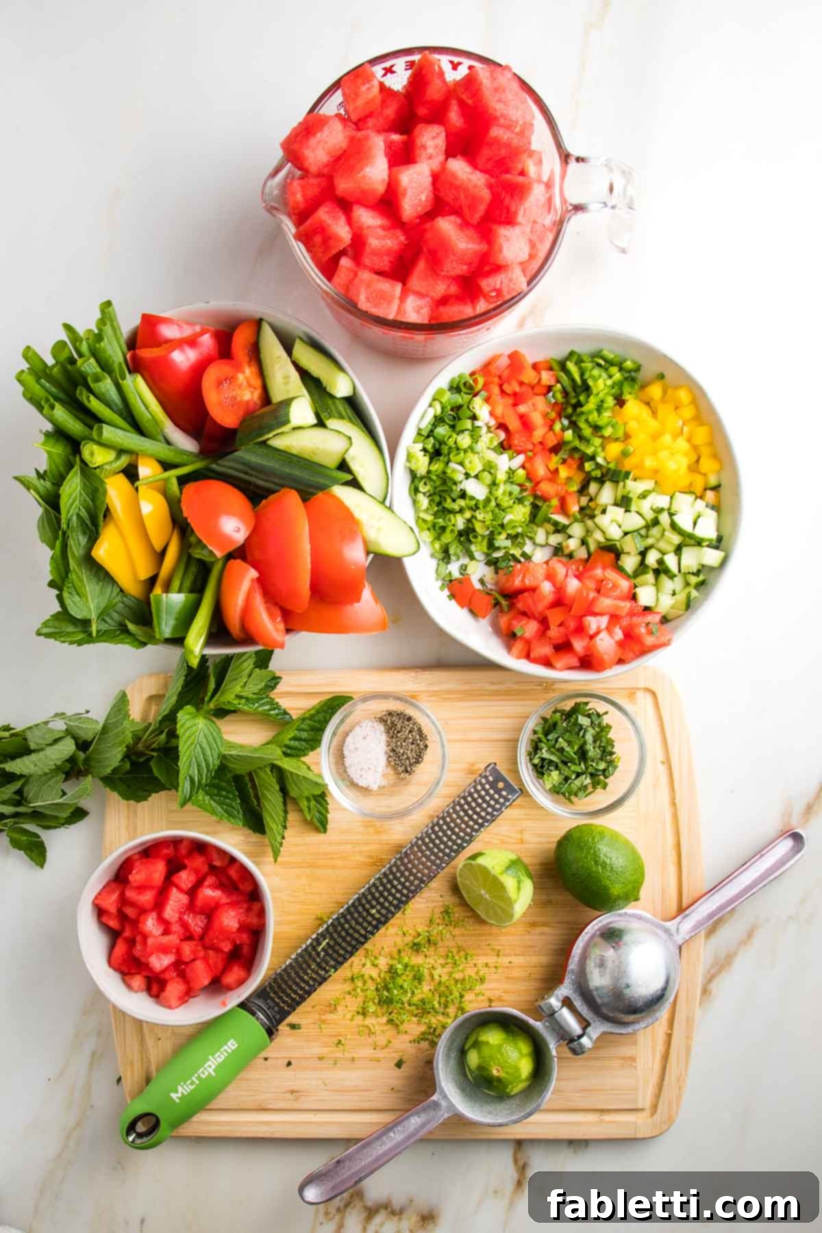 Everything is meticulously prepped and ready to create Watermelon Gazpacho. A bowl holds perfectly cubed watermelon, another contains chunky vegetables to be pureed for the soup, and a third bowl features small diced vegetables for a refreshing topping. Fresh mint leaves, a zested lime, a juicer, and seasonings like salt and pepper complete the vibrant mise en place.