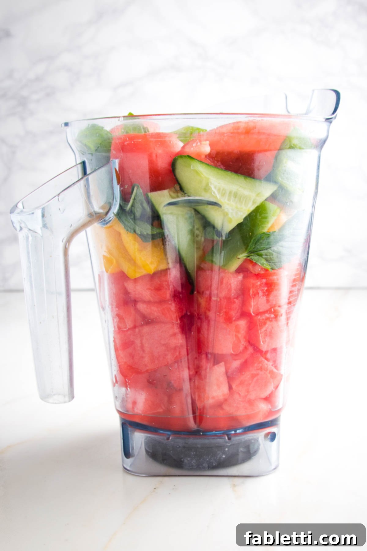 A close-up view of a high-speed blender pitcher, brimming with generous chunks of watermelon, vibrant red and yellow peppers, crisp cucumbers, ripe tomatoes, slender scallions, and fragrant fresh mint leaves. The ingredients are perfectly poised to be blended into a smooth and refreshing gazpacho.