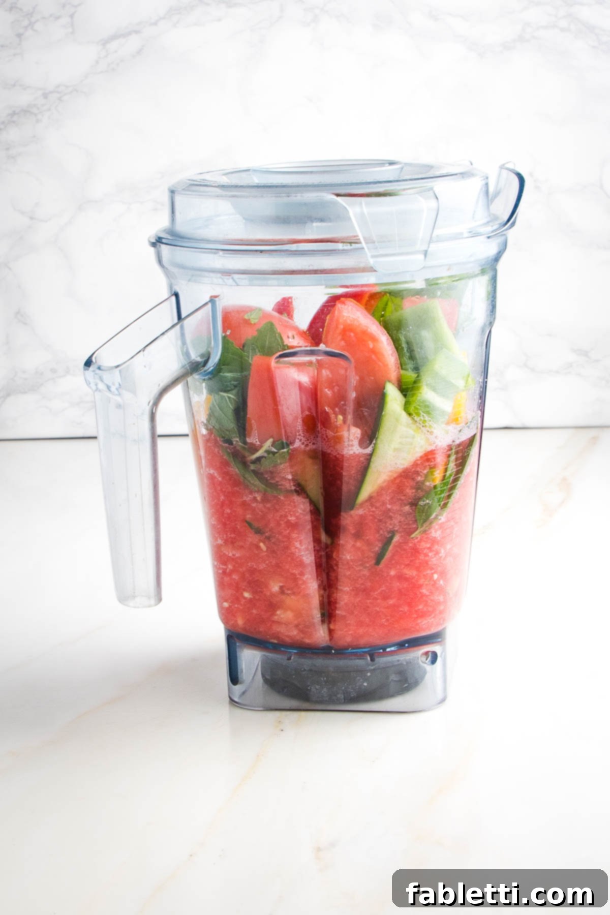 A high-speed blender pitcher captures the mesmerizing process of gazpacho creation. The bottom portion is a perfectly blended, smooth, pinkish-red liquid, while the top reveals ingredients yet to be incorporated—chunks of fresh tomato, crisp cucumber, sweet watermelon, and fragrant mint leaves, awaiting their turn.