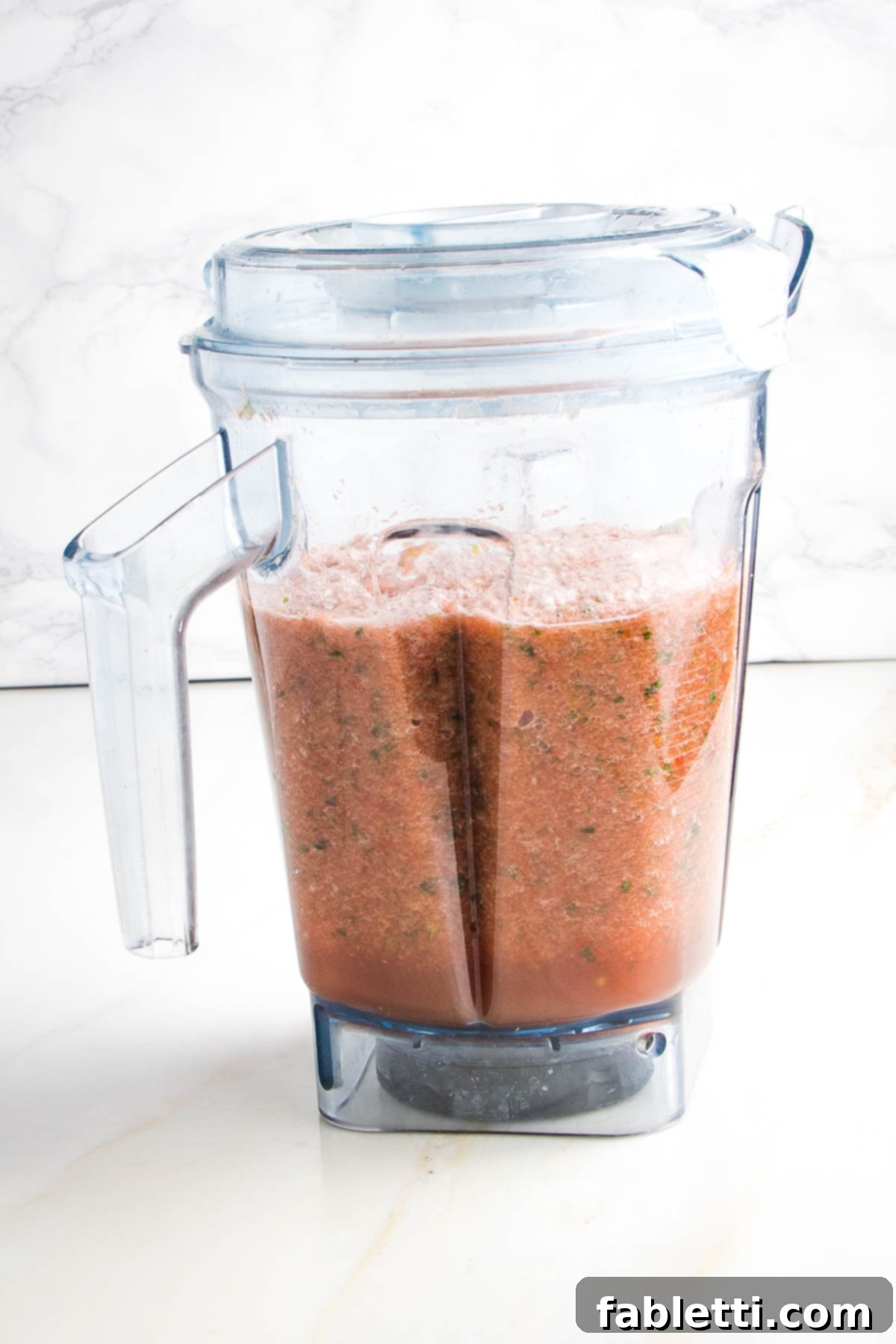 The high-speed blender pitcher now holds a perfectly smooth and deeply red Watermelon Gazpacho, with only tiny, vibrant flecks of green from the fresh herbs remaining, indicating a thoroughly blended, refreshing concoction.