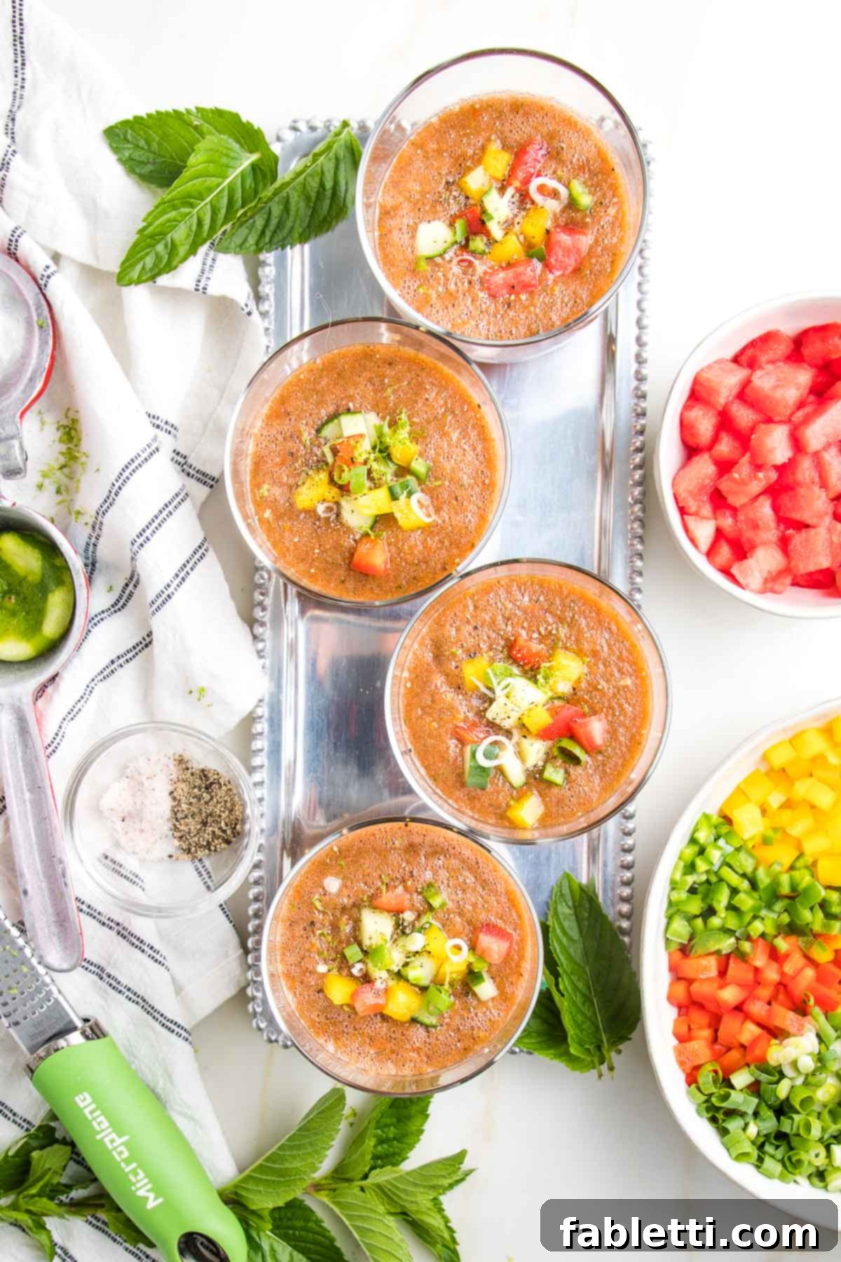Four elegant glasses of Watermelon Gazpacho are beautifully presented, each adorned with vibrant chunks of colorful diced vegetables floating atop. Alongside, a bowl of extra cut-up veggies and a bowl of fresh watermelon chunks are ready for garnish. A white towel with blue stripes, bright lime zest and wedges, and fragrant fresh mint leaves complete the refreshing display.