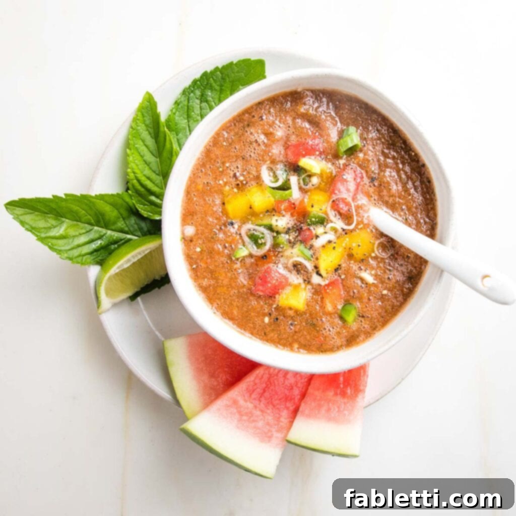 A pristine white bowl overflows with luscious watermelon gazpacho, generously topped with an abundance of finely diced, vibrant vegetables. The bowl rests on a rustic plate, elegantly garnished with sprigs of fresh mint, a bright lime slice, and several charming watermelon triangles.