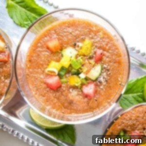 A close-up of a glass filled with watermelon gazpacho with small diced, colorful veggies and cracked black pepper. Glass is on a silver tray with other glasses of gazpacho and also some mint leaves and slices of lime.