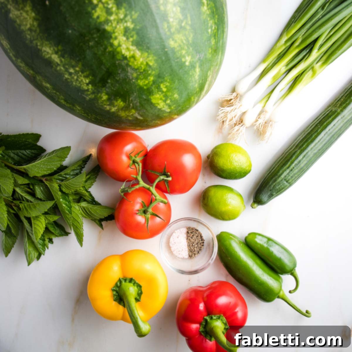 A vibrant arrangement of Watermelon Gazpacho ingredients: crisp watermelon chunks, fragrant fresh mint leaves, slender scallions, cool cucumber, ripe fresh tomatoes, bright limes, colorful red and yellow bell peppers, fiery jalapeños, and a sprinkle of salt and pepper.