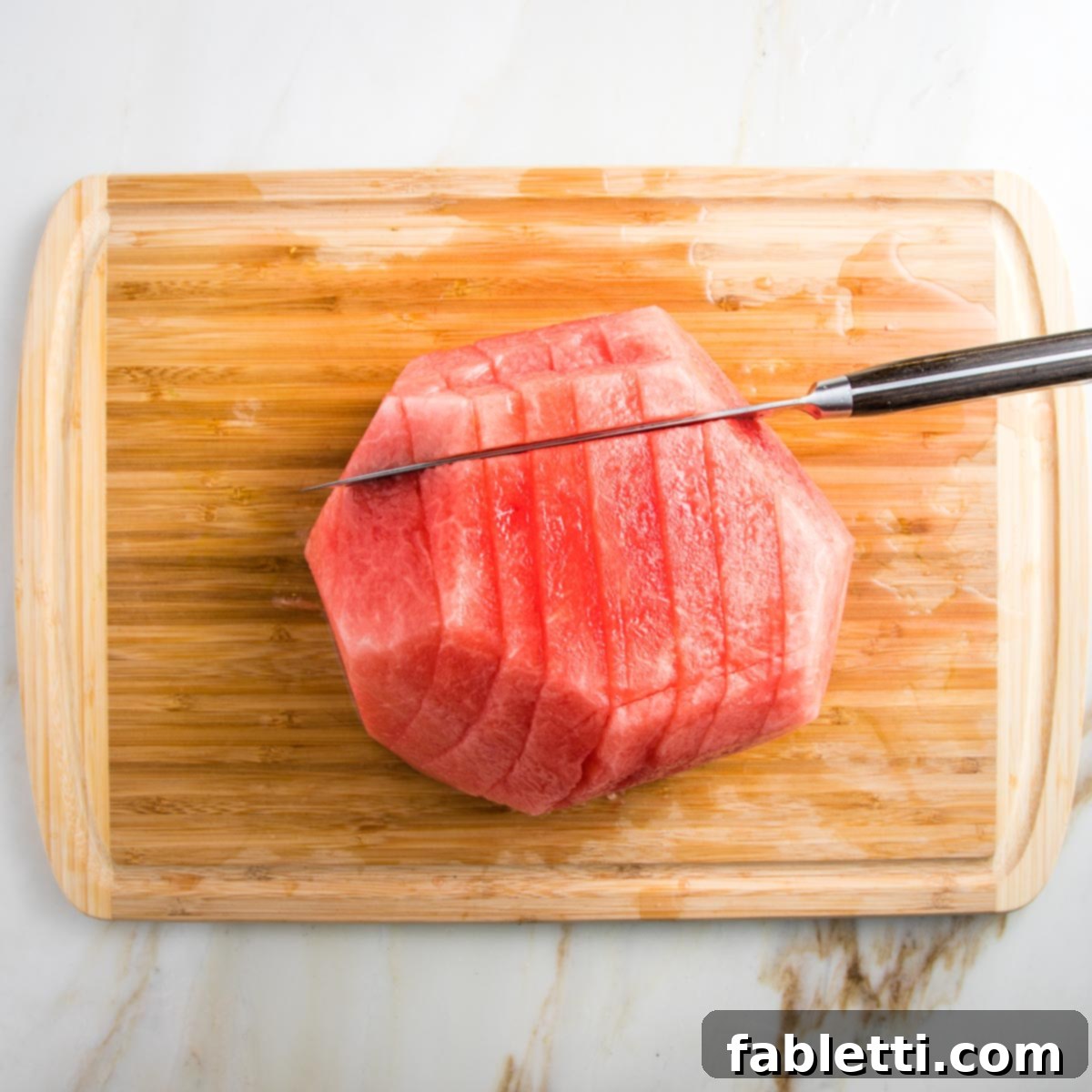 A wooden cutting board holds half a watermelon, its rind already removed. The melon is expertly sliced lengthwise and now being cut in the opposite direction, creating perfect spears.