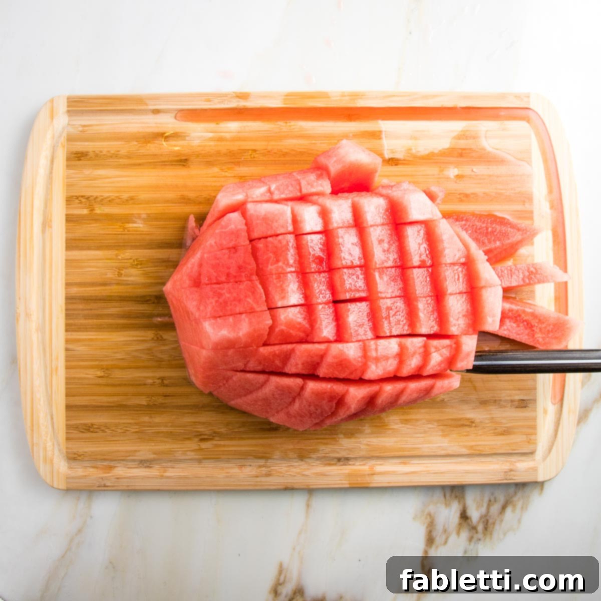 A close-up of a wooden board demonstrates the technique for cubing watermelon, with the board's natural groove expertly catching the delicious watermelon juice as it's cut.