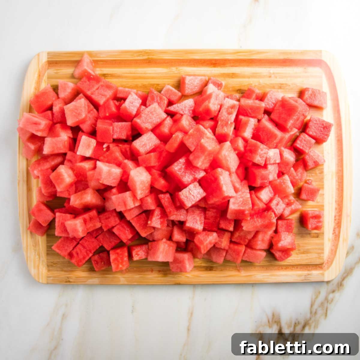 A wooden cutting board is laden with perfectly cut chunks of vibrant watermelon, with its luscious juice pooling in the board's convenient groove, ready for blending.