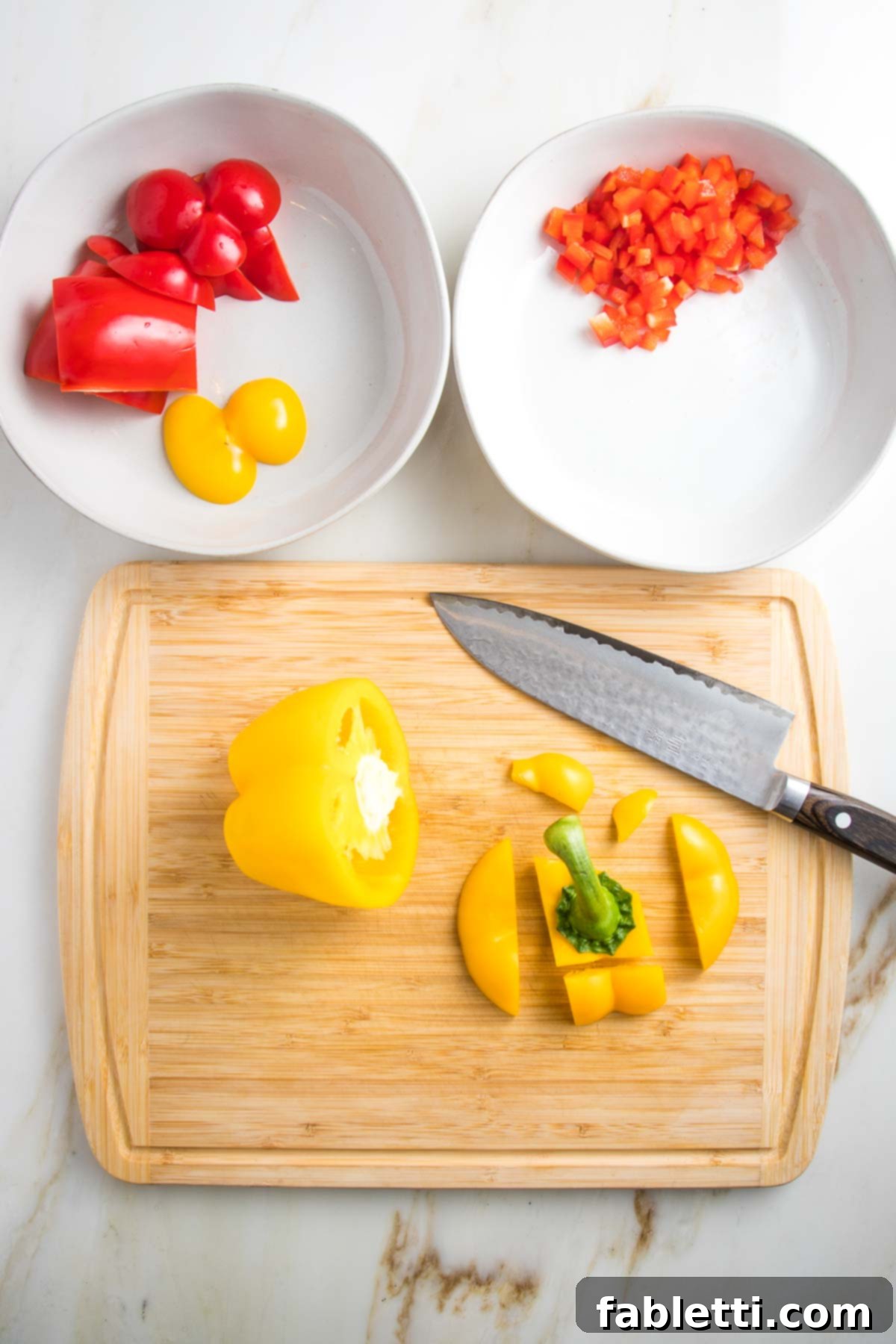 The image shows vegetable preparation in progress: two white bowls, one filled with chunky red and yellow bell peppers for blending, and the other with small diced red peppers for garnish. A yellow bell pepper is being diced on the cutting board.