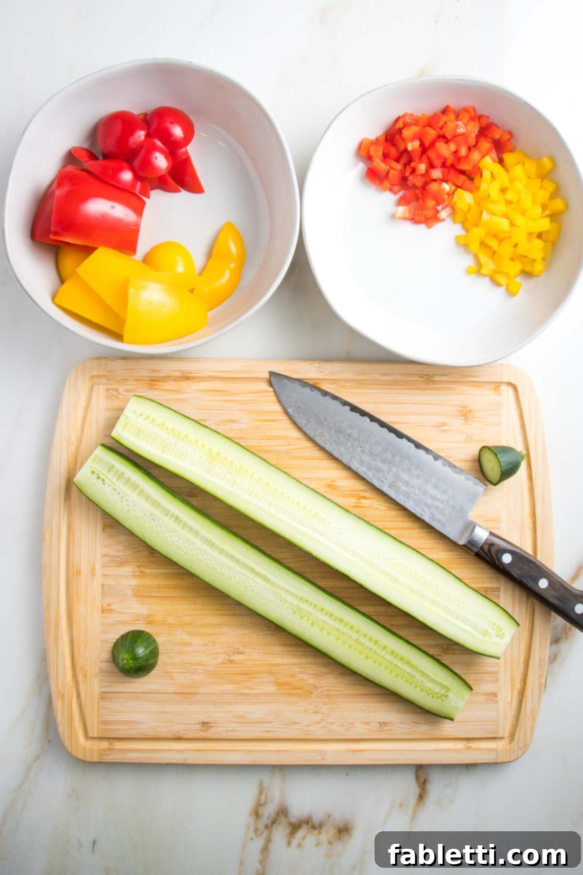 Two white bowls are shown: one with rough-chopped bell peppers, the other with minced yellow and red peppers. A bamboo cutting board features a halved English cucumber and a chef's knife, ready for preparation.