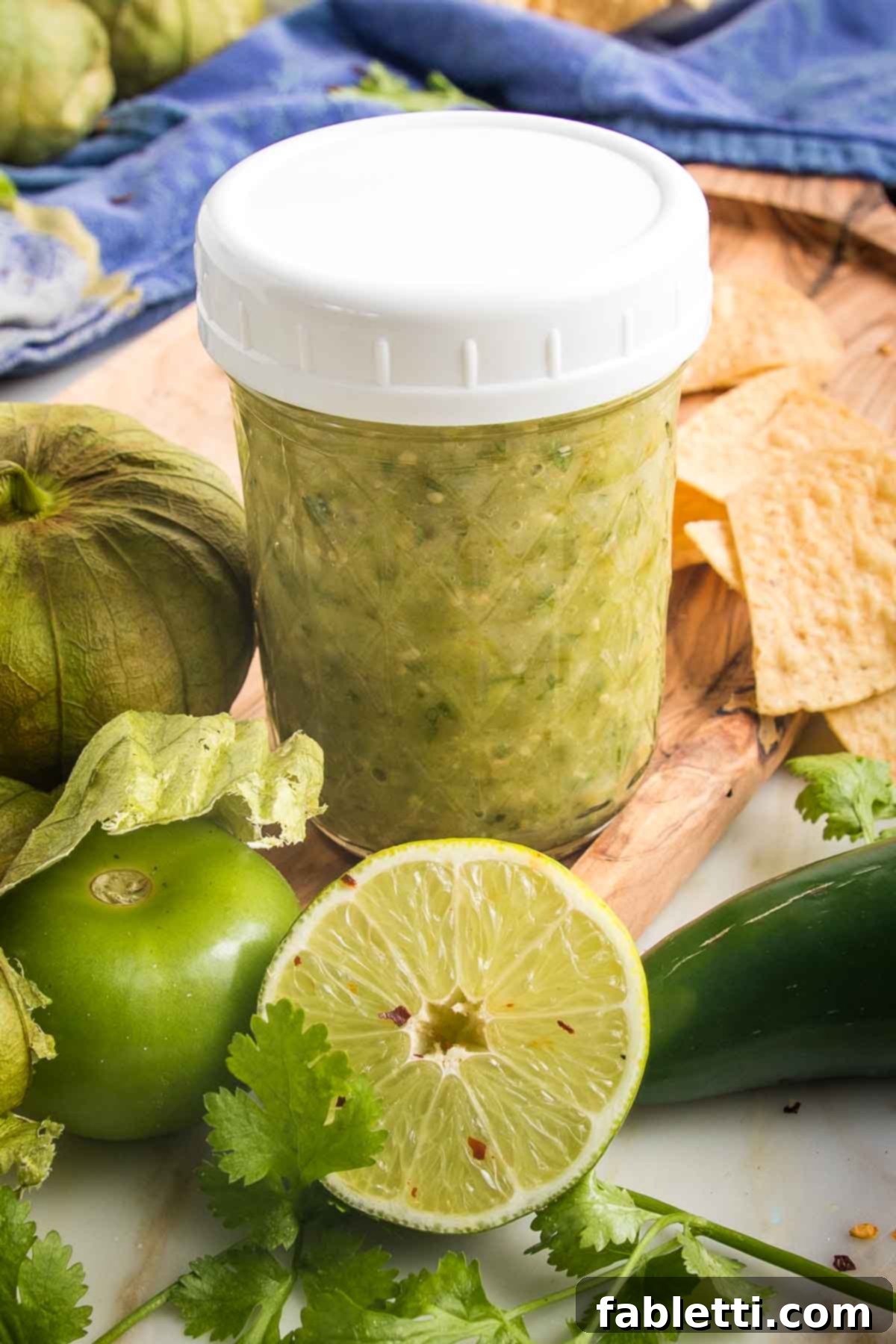 Fire-Kissed Tomatillo Salsa Verde 13 Small mason jar filled with green salsa, surrounded by fresh tomatillos, half a lime, fresh cilantro and tortilla chips.
