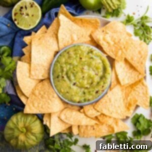 Fire-Kissed Tomatillo Salsa Verde 14 Green tomatillo salsa verde in a small bowl, surrounded by tortilla chips with a lime, a fresh tomatillo and some cilantro surrounding.