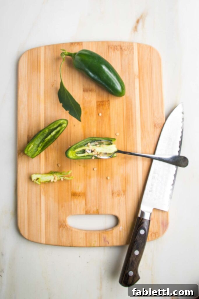 Fire-Kissed Tomatillo Salsa Verde 5 Small cutting board with one whole jalapeno and one sliced in half lengthwise, showing a grapefruit spoon scooping out the membranes and seeds.