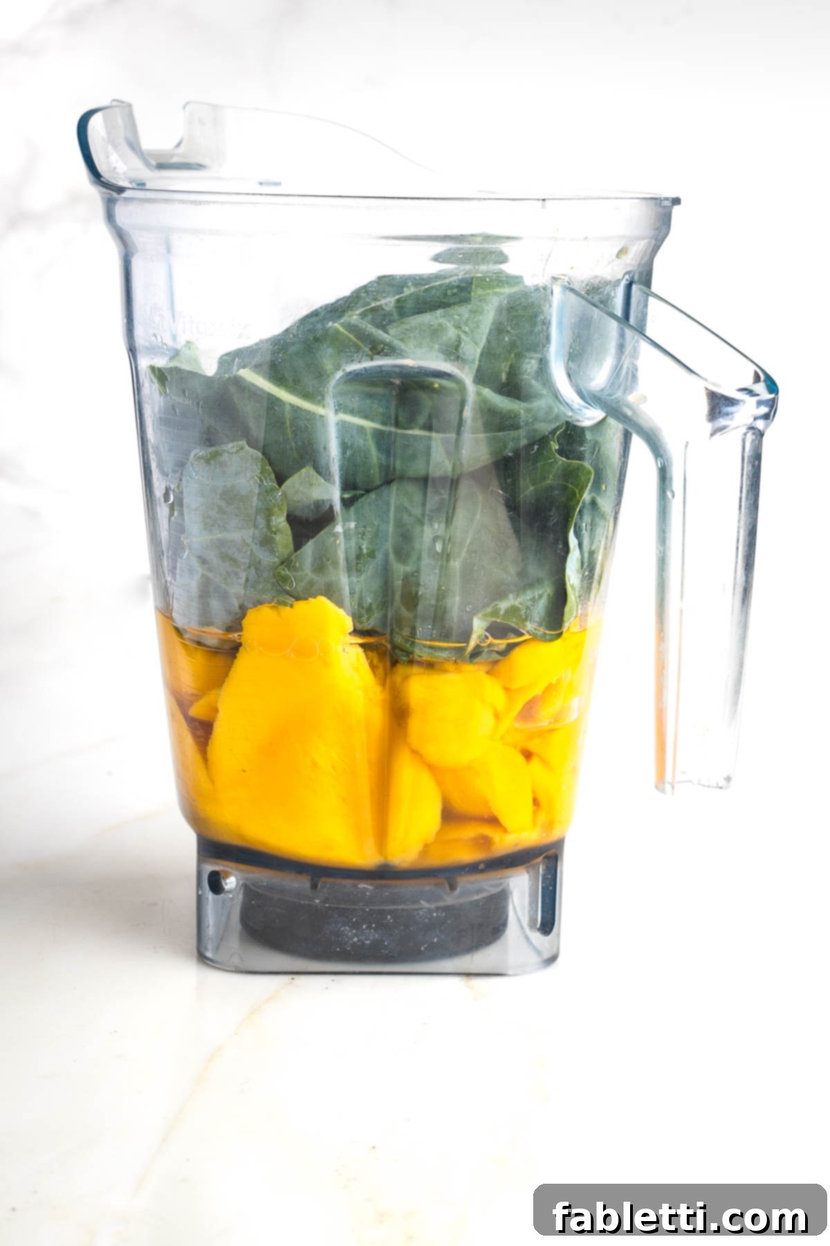 Blender pitcher with orange mango chunks, collard green leaves and water.