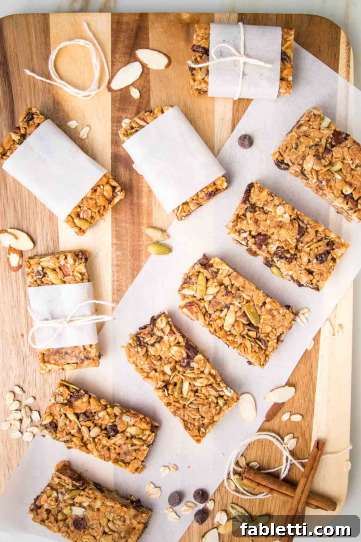 Golden Baked Granola Bites 2 Wooden board with granola bars lined up on a piece of parchment. Also are some bars wrapped in parchment and tied with a kitchen twine. Some cinnamon stick and loose nuts and chocolate chips are strewn about.