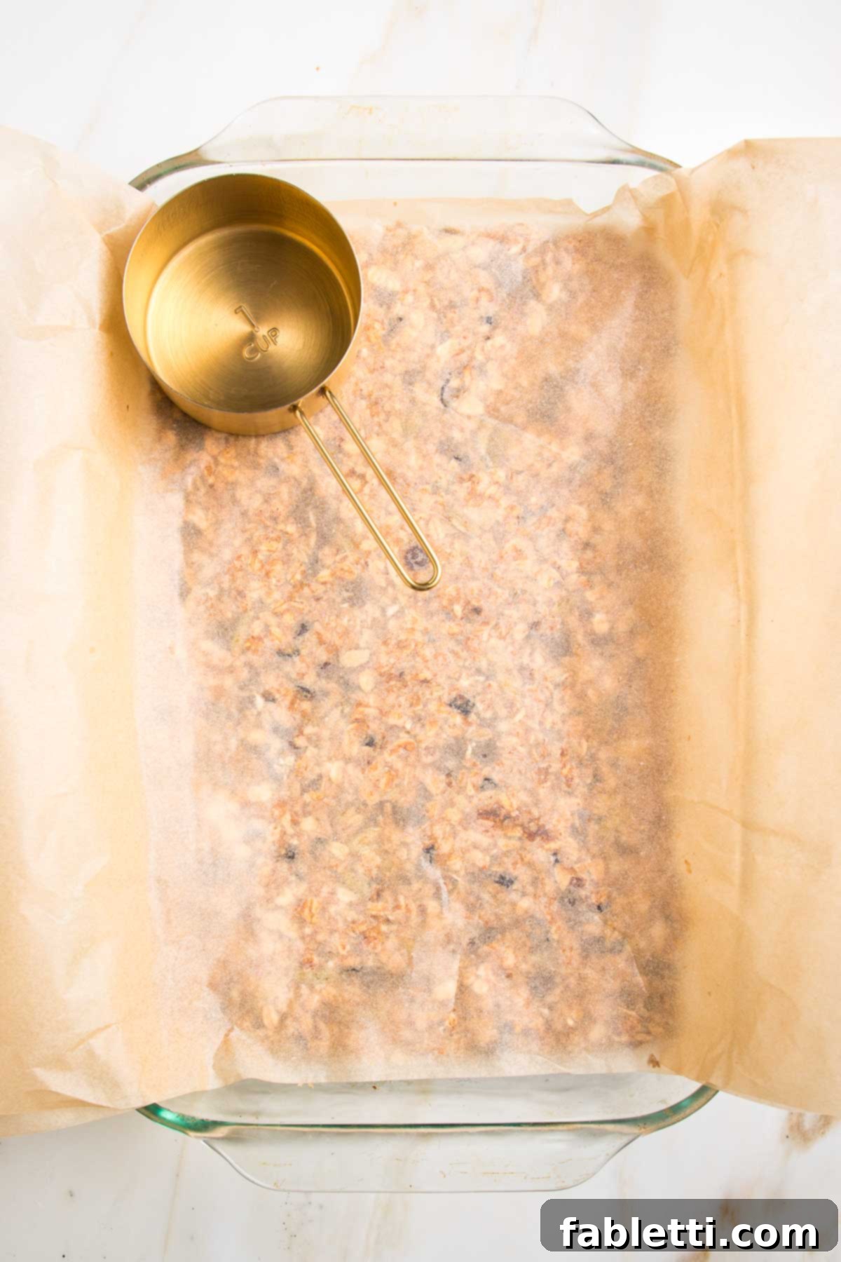 Golden Baked Granola Bites 14 Glass baking dish filled with granola bar mixture, covered with another sheet of parchment, and being pressed down firmly with a gold measuring cup.