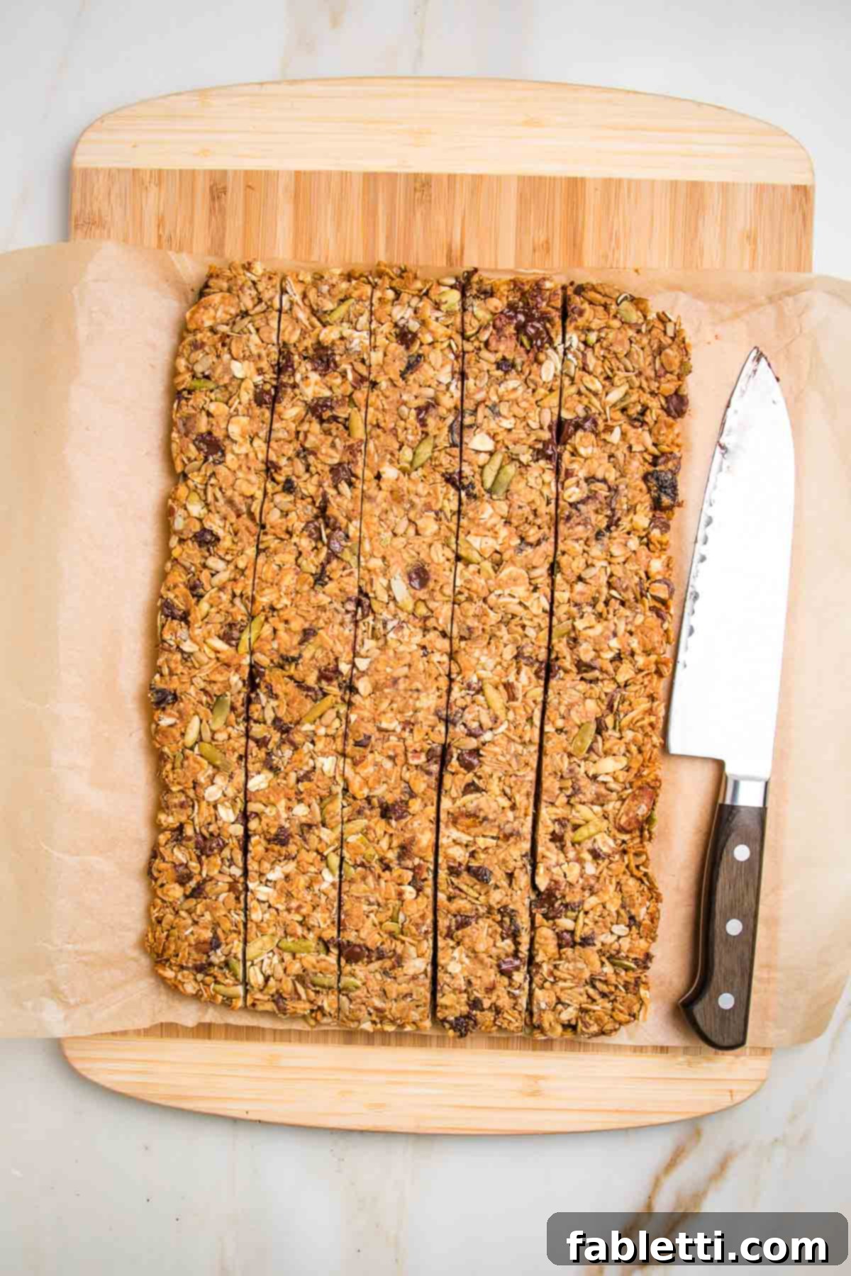 Golden Baked Granola Bites 16 Granola bars on a piece of parchment atop a cutting board, cut into long thin strips with a sharp knife resting on the side.