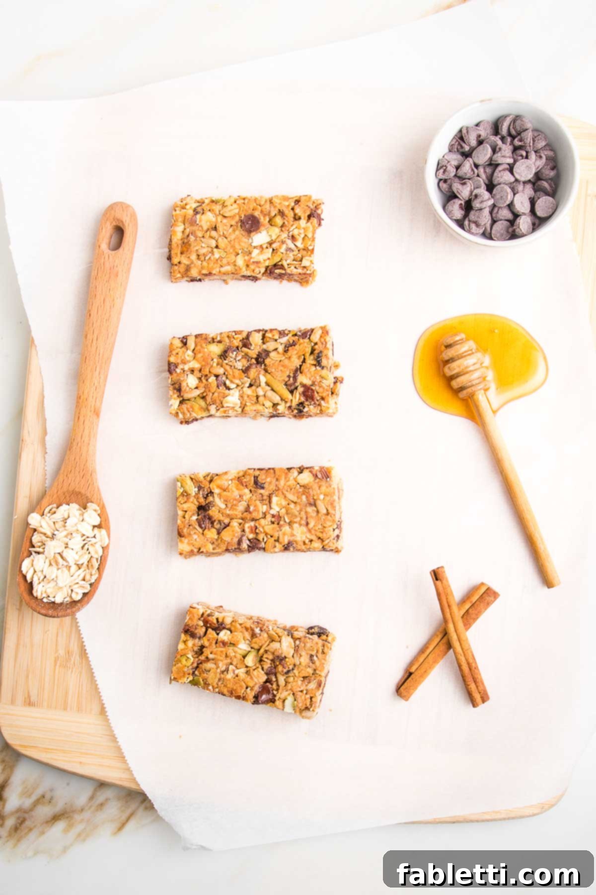 Golden Baked Granola Bites 20 4 crunchy granola bars lined up on a piece of parchment. There is a spoon full of oats, a puddle of honey, a dish of chocolate chips and two cinnamon sticks.