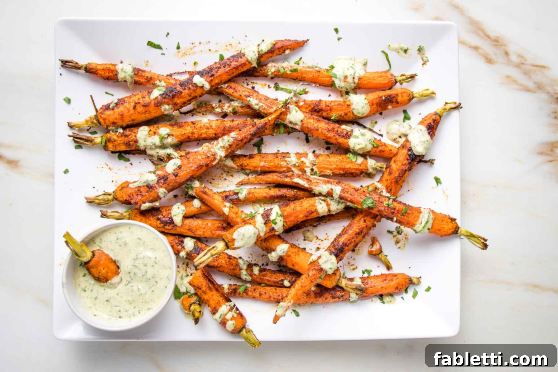 White platter of whole roasted carrots, drizzled with tahini sauce and a small dish of sauce on the side for dipping.
