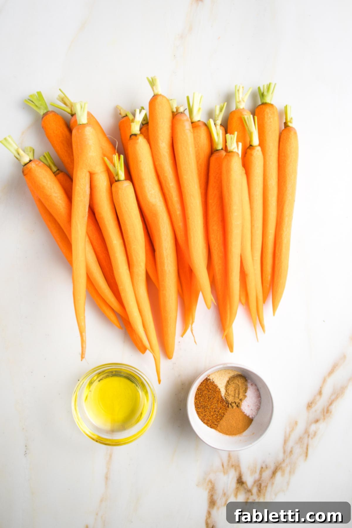 Whole, peeled orange carrots on a white counter with a small dish of olive oil and another dish with an array of dried spices. 