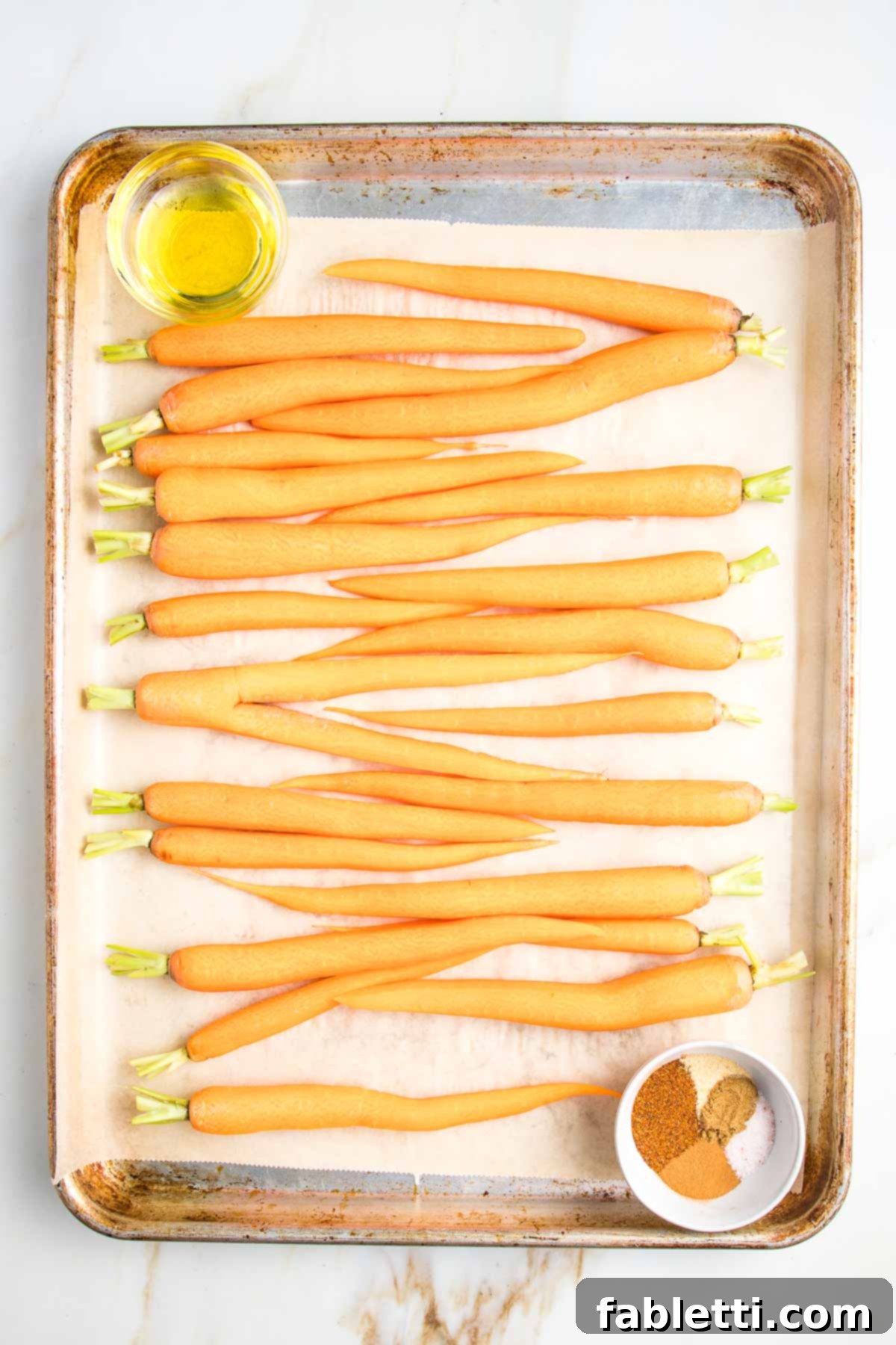 Parchment lined baking tray with whole, trimmed carrots, a small dish of olive oil and another small dish with spices.