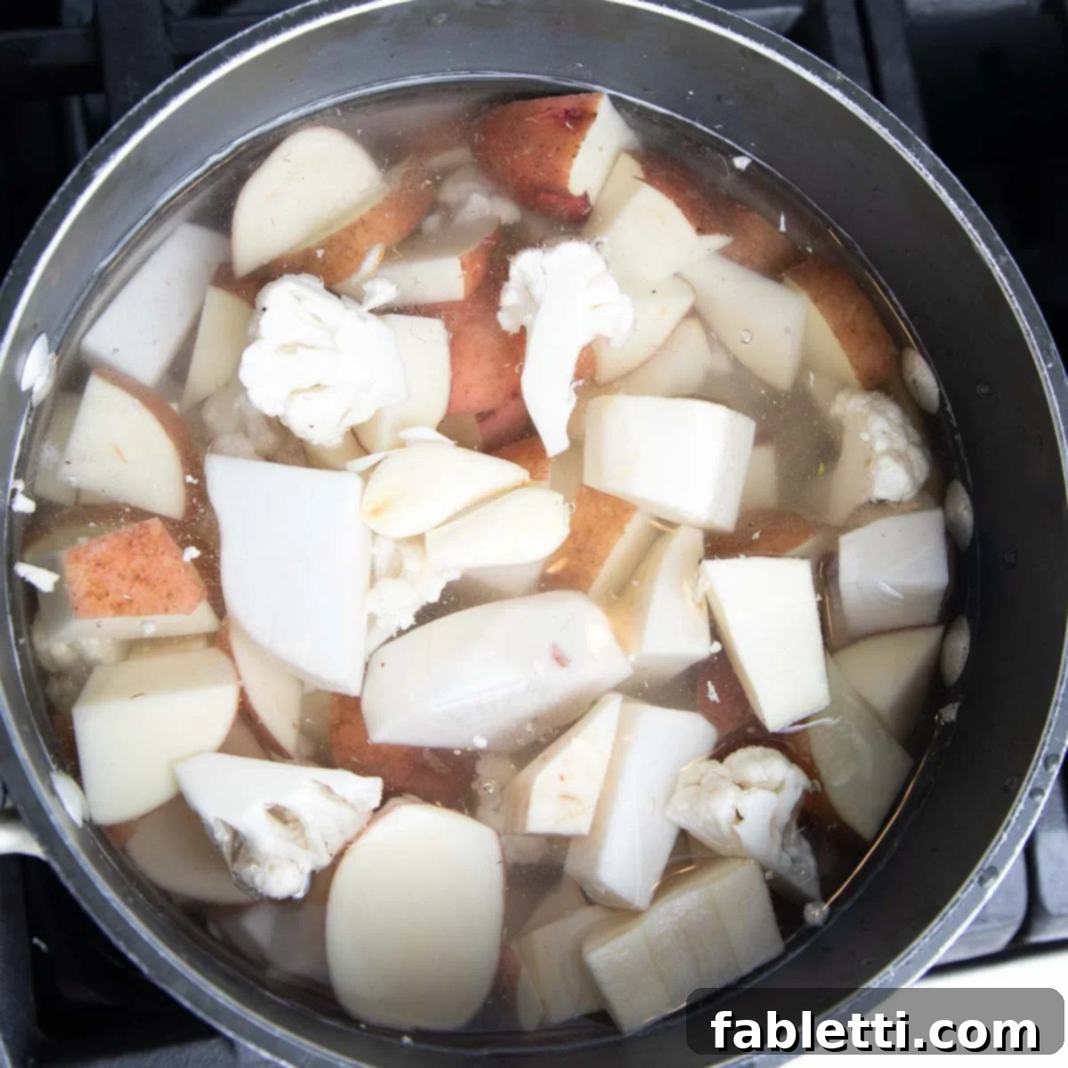 Heavenly Dairy-Free Garlic Mash 5 A pot on the stove, filled with cubed root vegetables, cauliflower, and whole garlic cloves, covered with water, brought to a simmer.