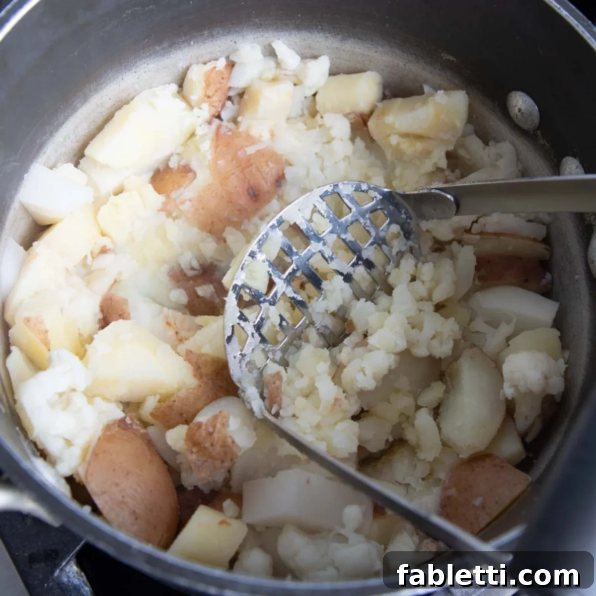 Heavenly Dairy-Free Garlic Mash 7 A potato masher in action, mashing cooked root vegetables, cauliflower, and garlic directly in the pot, creating a creamy texture.
