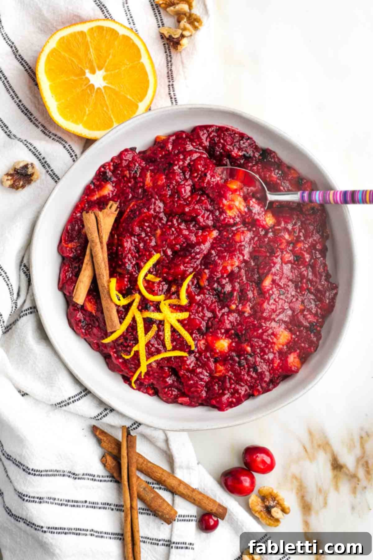 Refined Sugar-Free Cranberry Sauce 2 A rustic bowl brimming with chunky homemade cranberry sauce, garnished with vibrant orange zest and a sprinkle of toasted walnuts. Cinnamon sticks and fresh cranberries artfully arranged around the bowl hint at the delicious flavors within.