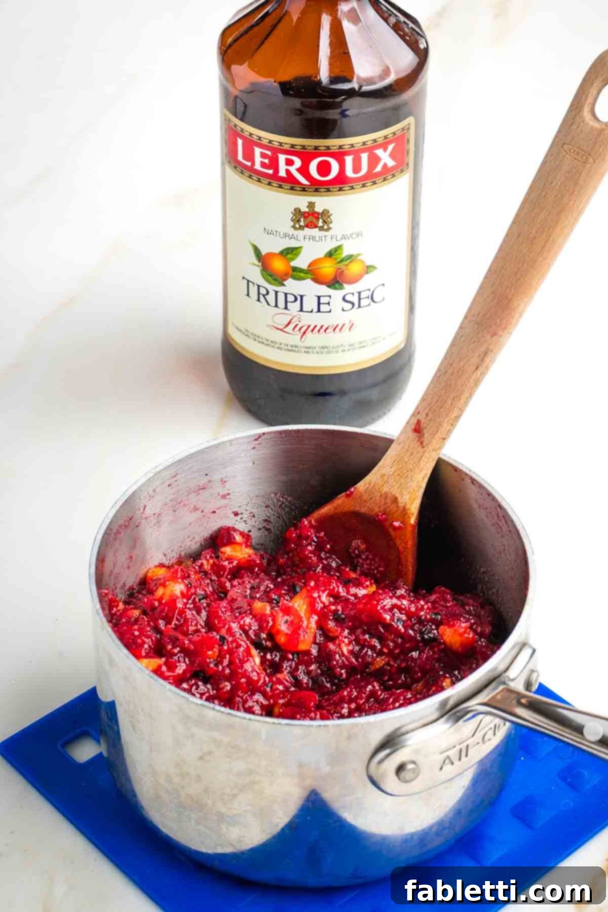 Refined Sugar-Free Cranberry Sauce 11 A hand gently stirring a pot of freshly made cranberry sauce, with a small bottle of triple sec visible nearby, indicating the optional addition of the secret ingredient for enhanced flavor.