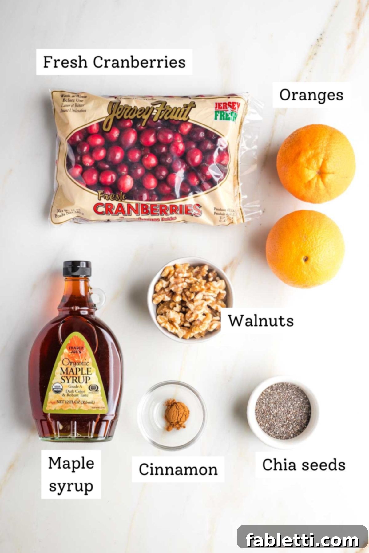 Refined Sugar-Free Cranberry Sauce 3 A beautifully arranged selection of fresh ingredients for cranberry sauce, including a bag of bright red cranberries, two whole oranges, a bottle of golden maple syrup, a handful of walnuts, cinnamon sticks, and a small dish of chia seeds, all ready for cooking.