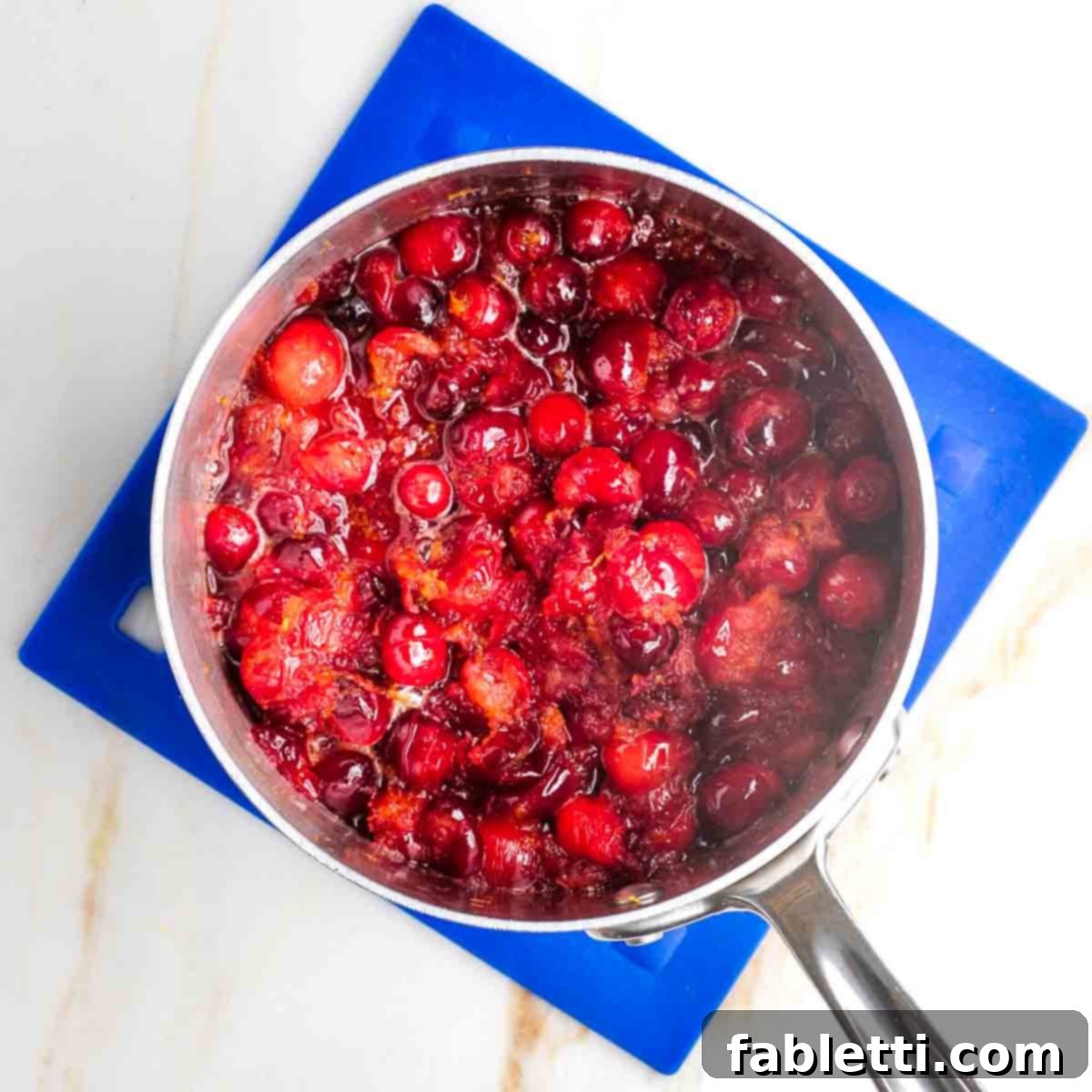 Refined Sugar-Free Cranberry Sauce 7 A close-up of a small pot on a stovetop, where fresh cranberries and orange pieces are gently simmering, their vibrant colors indicating the cooking process is well underway.