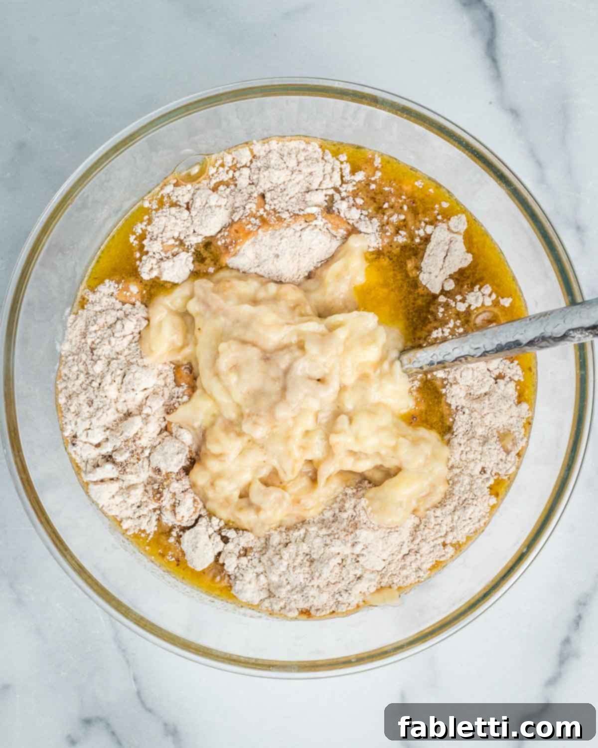 The wet ingredients, including mashed bananas, are poured into the bowl containing the dry ingredients, ready for mixing.