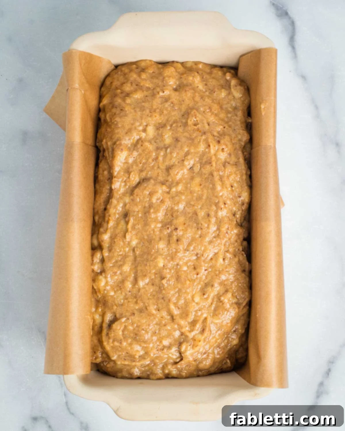 Banana bread batter poured into a loaf pan that is neatly lined with unbleached parchment paper, ready for baking.